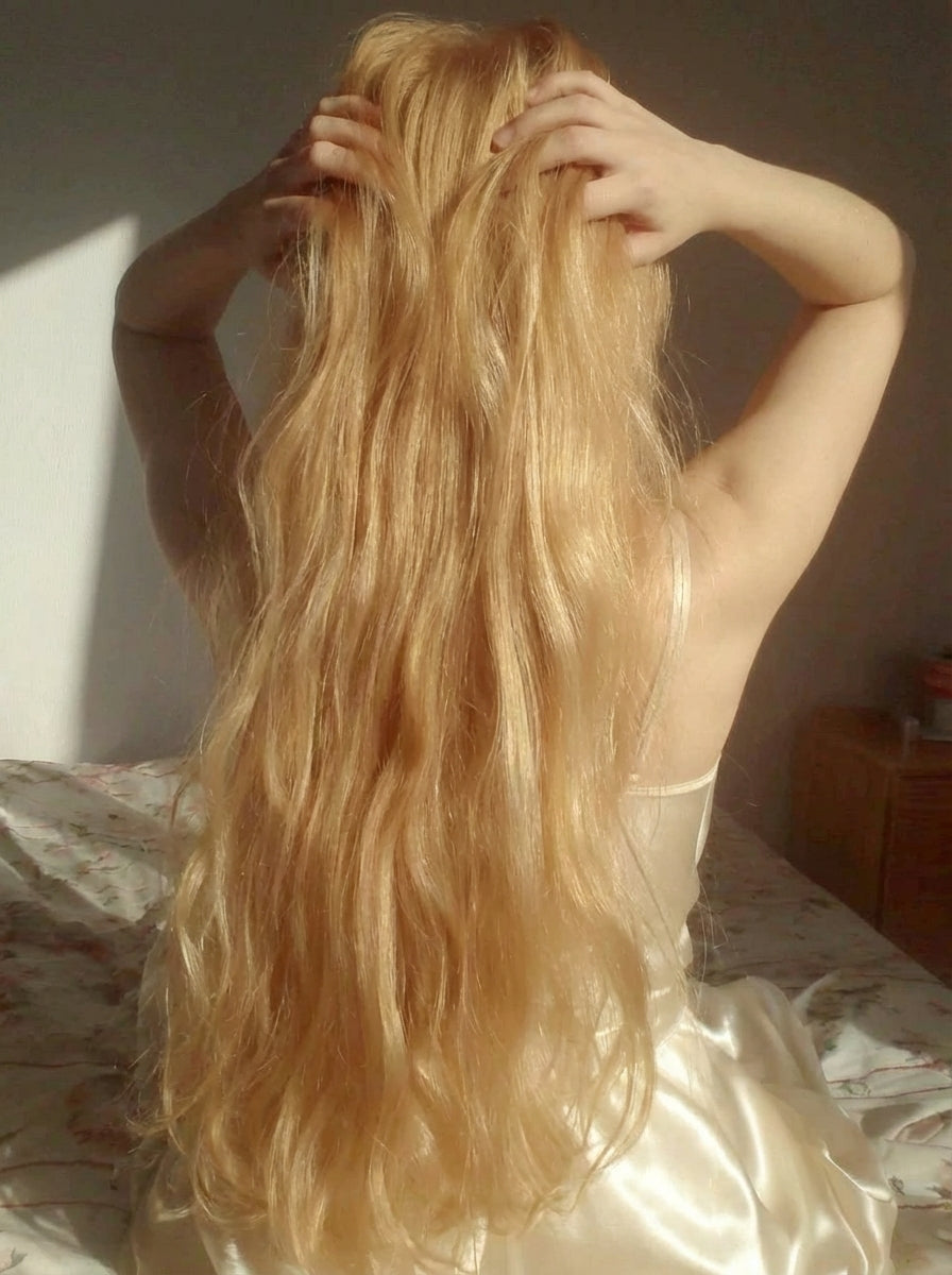 A woman seen from behind lifting her long blonde waves with both hands in sunlight.