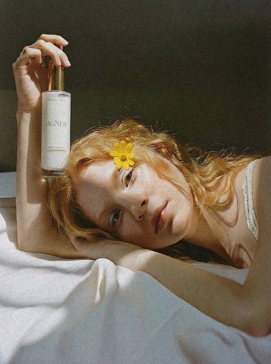 A red-haired woman lying on white linen with a yellow flower in her hair, holding the Agnes Hair Milk bottle.