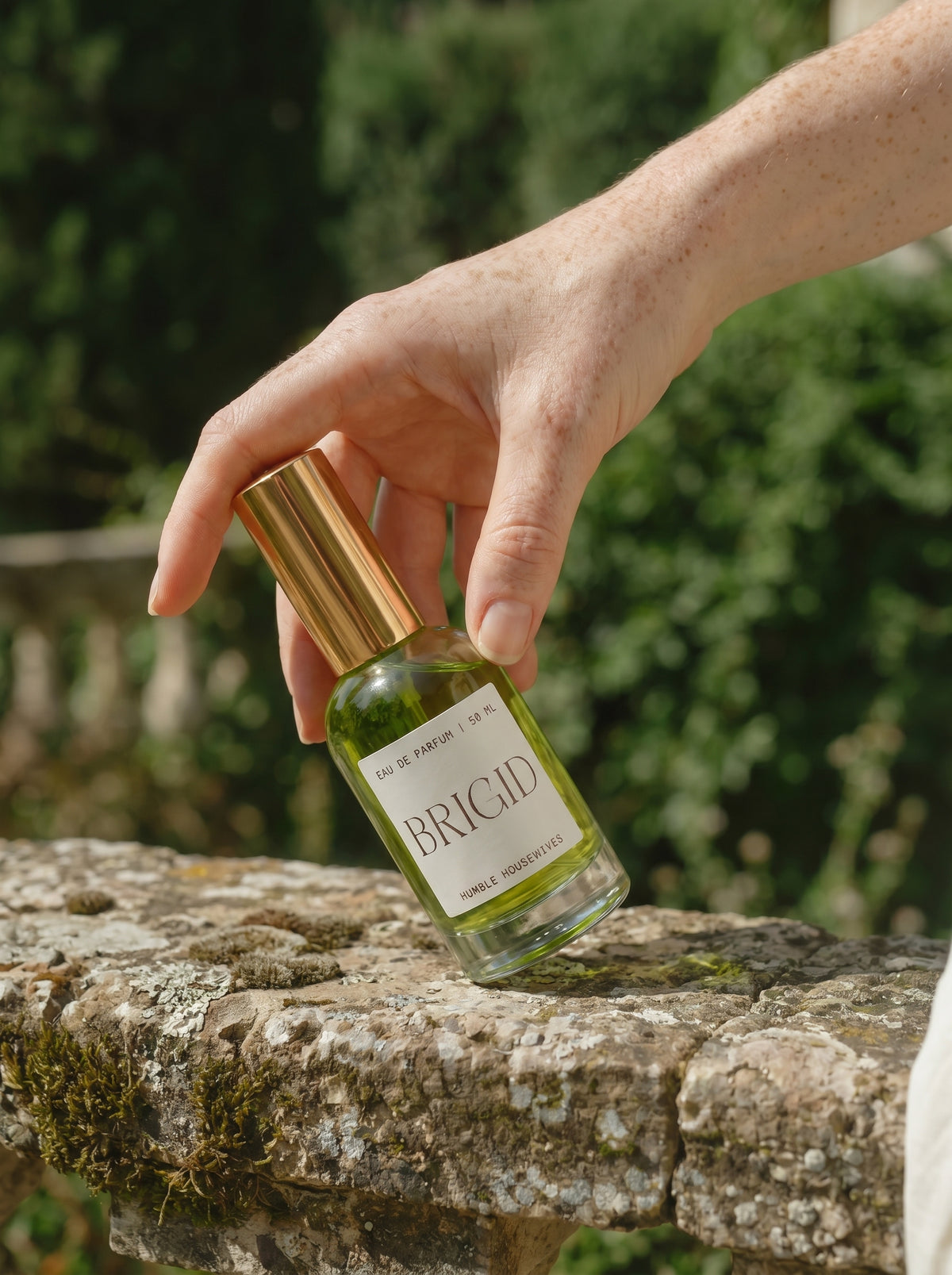 A hand setting the Brigid Eau de Parfum bottle on a mossy stone wall with greenery behind.