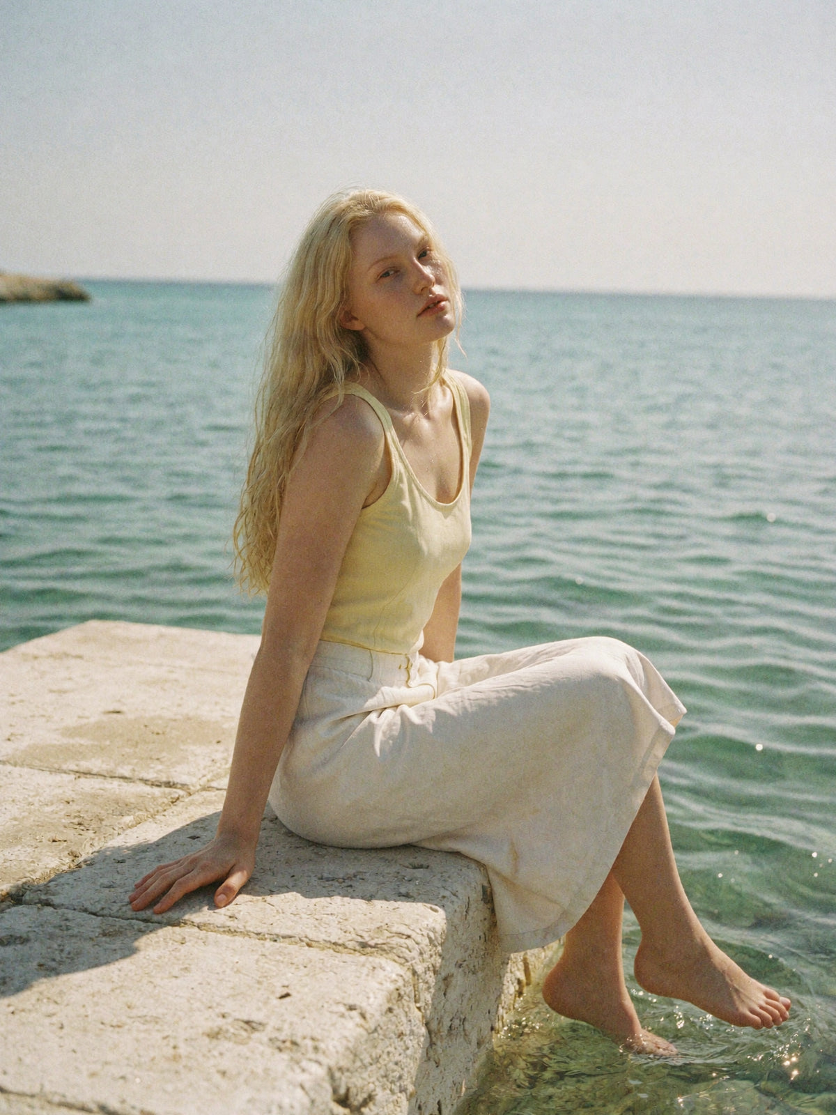A blonde woman in a yellow top sitting on a stone quay with her feet in clear turquoise water.